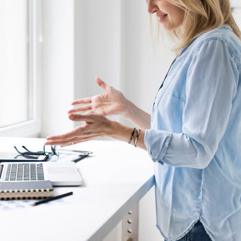 Blonde woman in an online conference call at a standing desk wearing a light blue shirt 