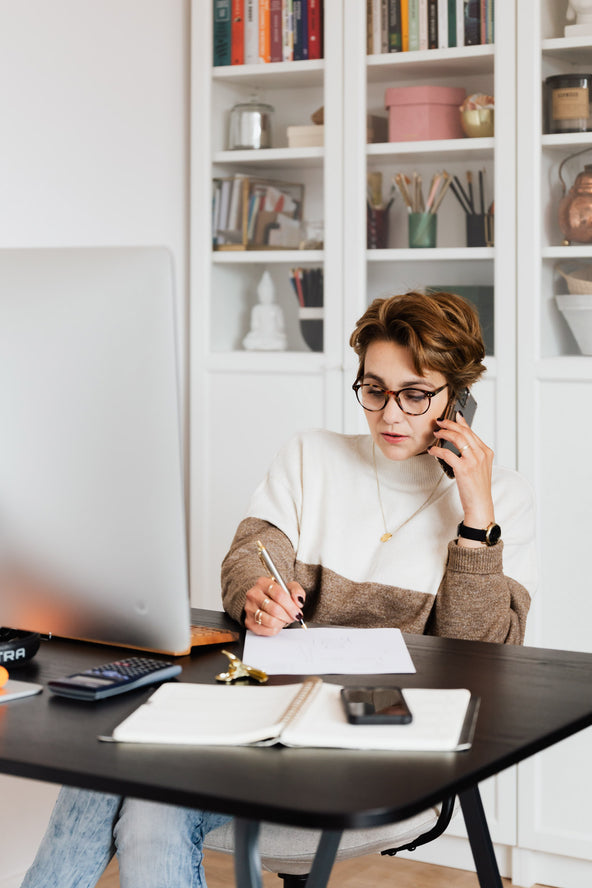 A woman sitting in her home office talking on the phone taking notes